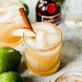 An apple cider margarita with ice, a cinnamon stick, and apple slice garnish in a rimmed glass, surrounded by limes and a liqueur bottle.