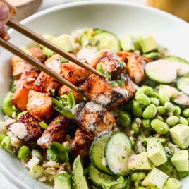 A bowl of bang bang salmon, avocado, edamame, cucumber, and rice, with chopsticks picking up a salmon piece.