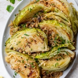 Braised cabbage wedges garnished with parsley and caramelized onions, served on an oval white plate. Lemon wedges and fresh parsley are visible on the side, set on a light-colored surface.