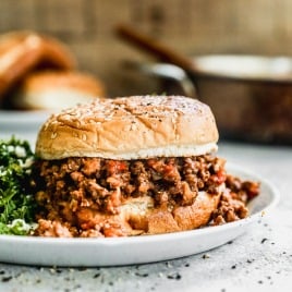 A sloppy joe sandwich with seasoned ground beef on a sesame bun, served on a white plate with a side of greens.