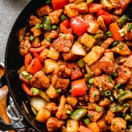 A skillet filled with stir-fried sweet and sour pork, bell peppers, onions, pineapple, and green onions.