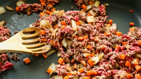 Chopped onions, carrots, and ground beef are being sautéed in a pan with a wooden slotted spoon, showing a mix of cooked and uncooked meat and vegetables.