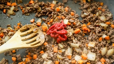 Ground beef, diced onions, and carrots are being cooked in a pan. A wooden spoon is mixing the ingredients, and a dollop of tomato paste sits on top of the mixture.