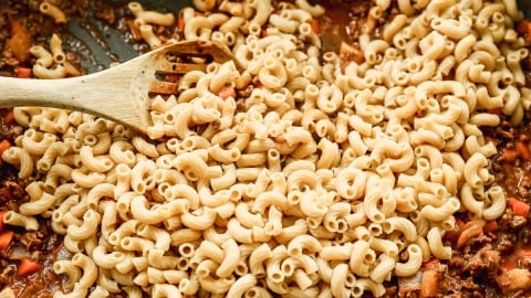 A wooden spoon rests on a skillet filled with cooked elbow macaroni, ground meat, red bell pepper pieces, and sauce, as the ingredients are being mixed together.