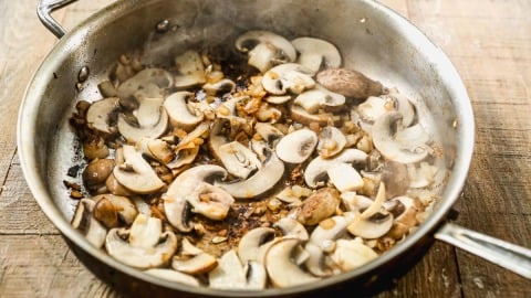 Sliced mushrooms and diced onions sautéing in a metal pan on a wooden surface, with visible steam rising.
