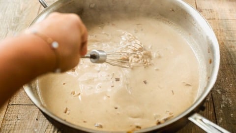 Hand whisking creamy sauce with onions in a large metal pan on a wooden table.