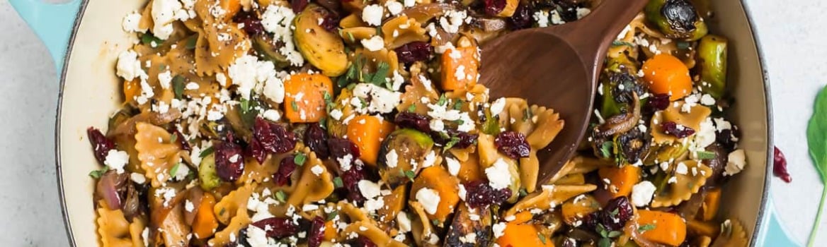 A skillet of pasta with butternut squash, cranberries, and cheese on a checkered towel, with a wooden spoon.